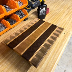 A striped end-grain cutting board with contrasting wood stripes and a bottle of cutting board oil beside it on a workbench