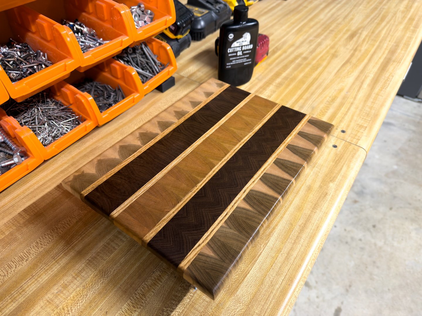 A striped end-grain cutting board with contrasting wood stripes and a bottle of cutting board oil beside it on a workbench