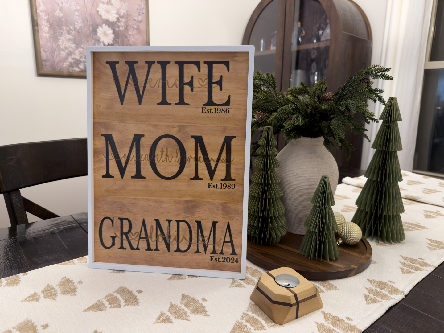 Personalized wood sign centerpiece for a family display A framed wooden sign reading wife, mom, and grandma sits on a decorated dining table with seasonal greenery and small tree ornaments.