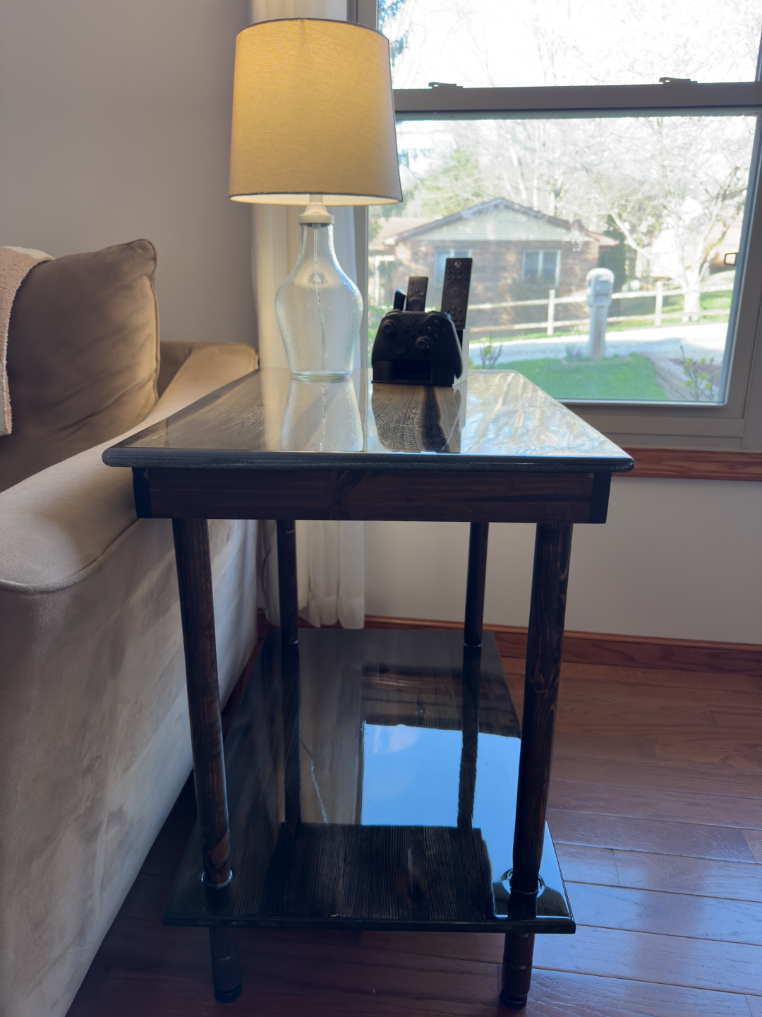Cozy living room side table with lamp by the window A wooden side table holds a table lamp and remote controls beside a sofa in a bright living room.