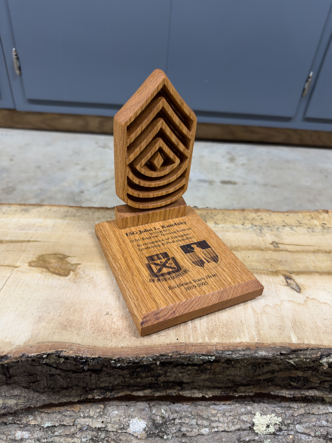 A wooden award plaque with three carved chevrons on an engraved base, standing on a log slab