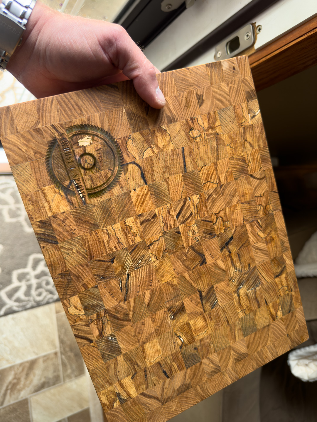 A hand holding a square end-grain cutting board with a wood-saw blade design burned into the grain