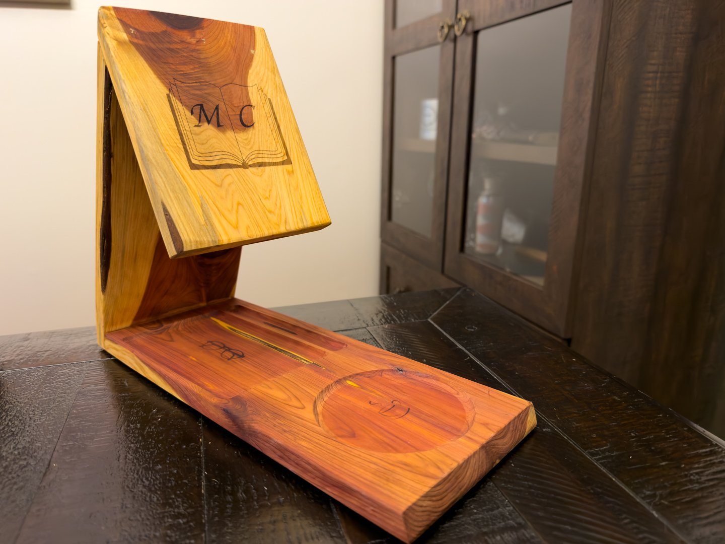 A cedar book, coffee, glasses, and pen holder on a dark table, with an open book on the angled rest