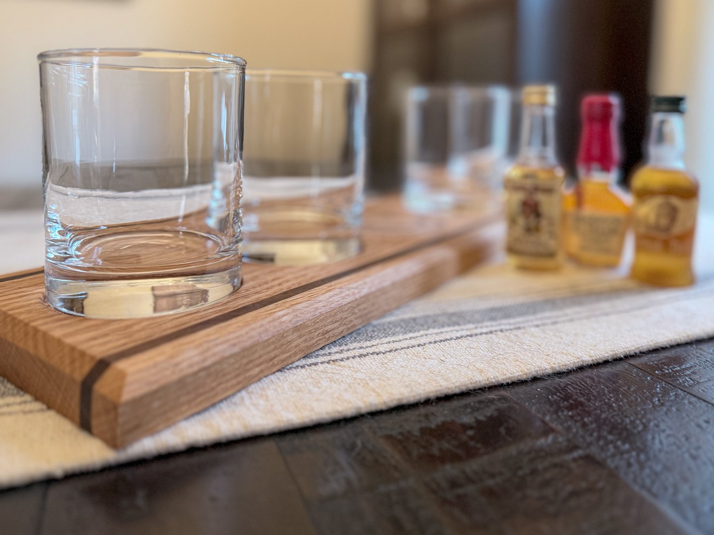 Four empty glass tumblers sitting in the recesses of a light oak whiskey flight board on a dark table.