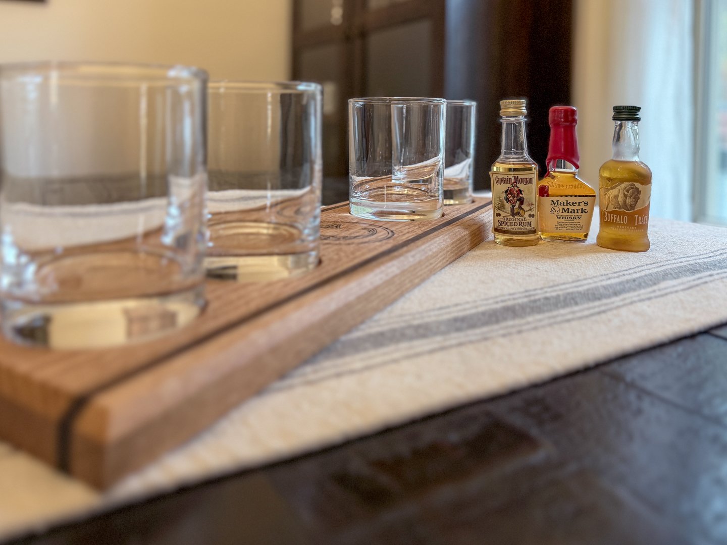 An angled view of a light oak whiskey flight board with glass recesses and sample bottles on a table.