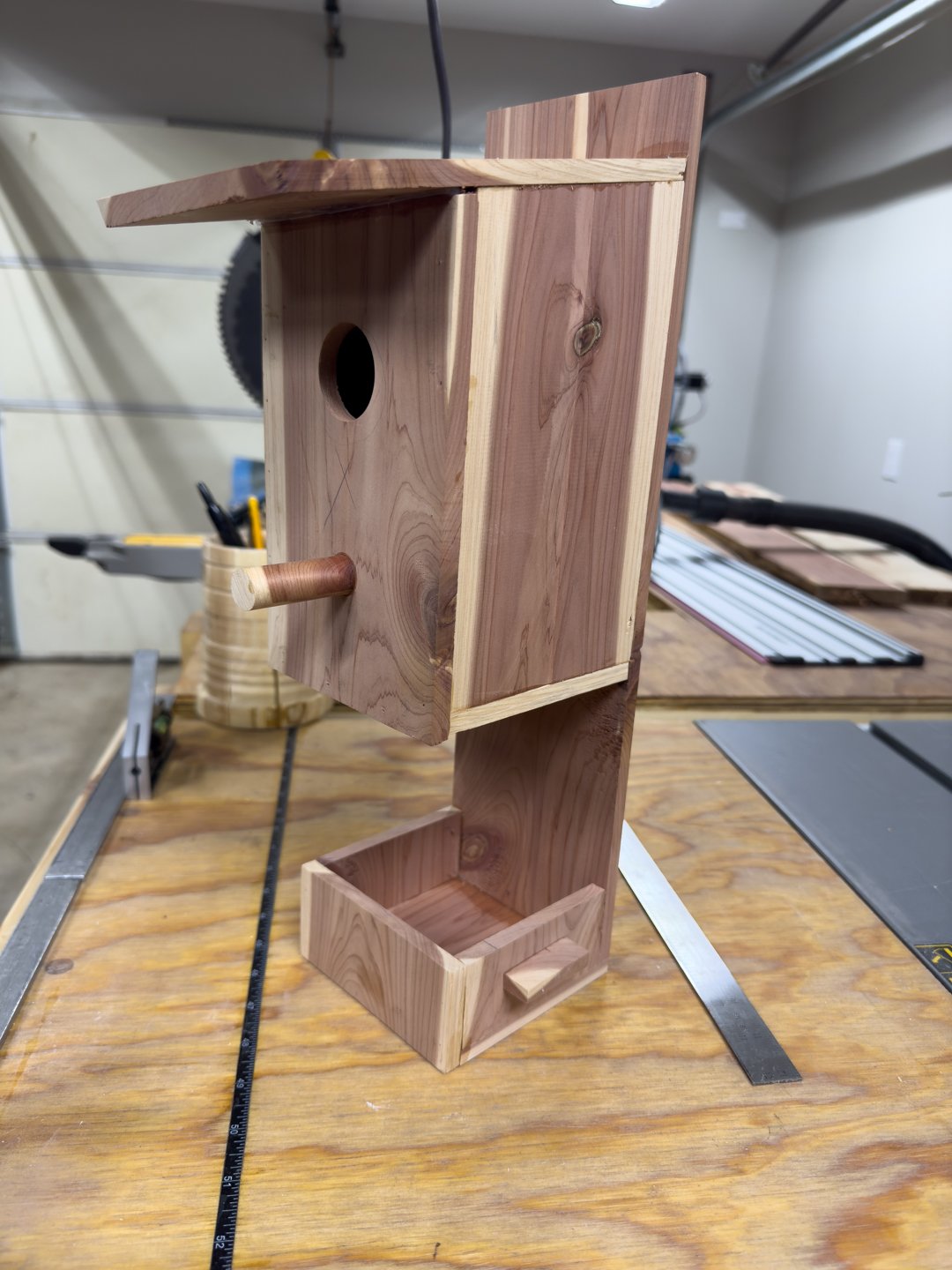 A cedar birdhouse in a workshop, showing the overhanging roof, entrance, and perch on a tall backboard.