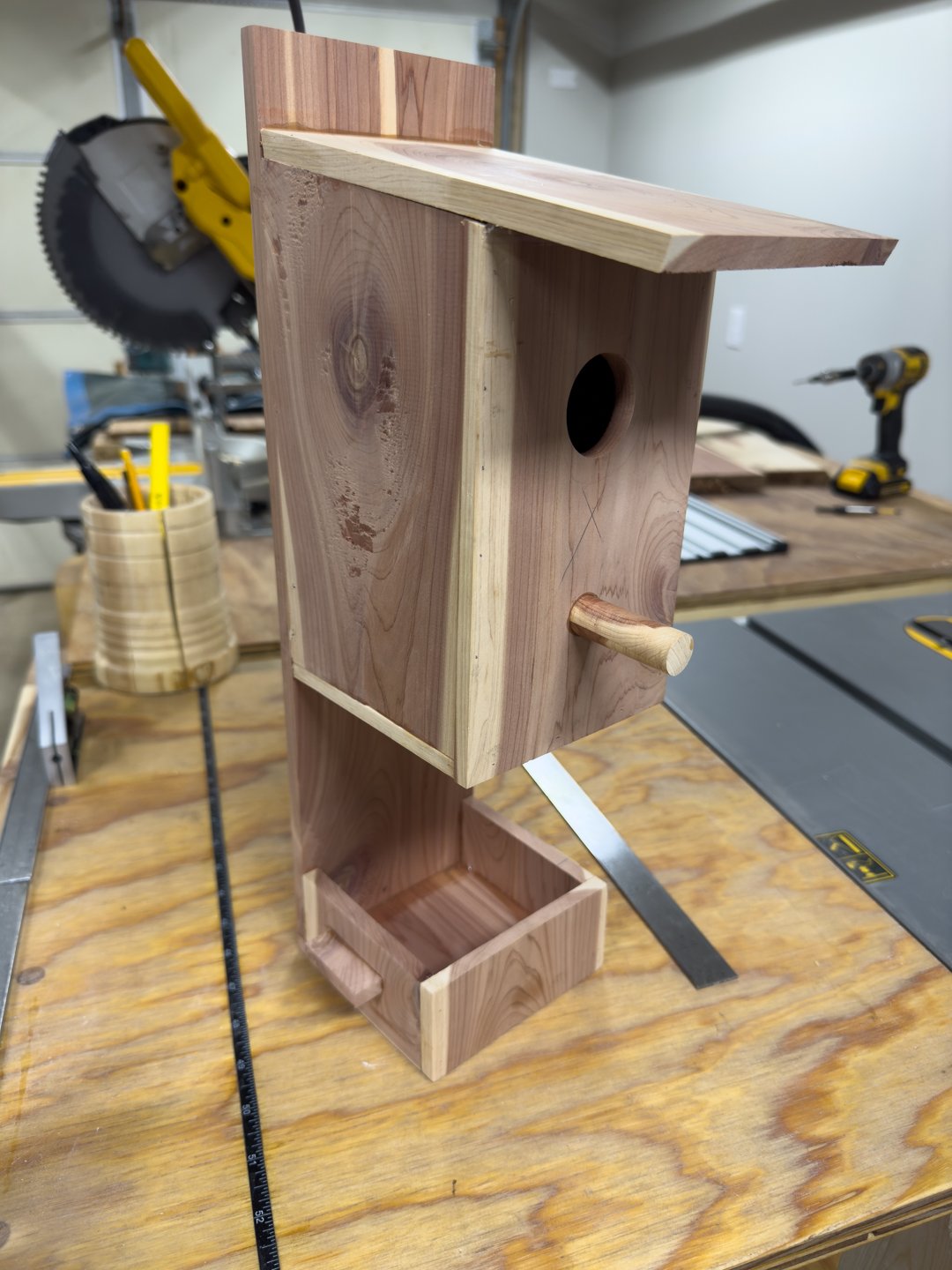 A cedar birdhouse with a feeding tray sitting on a workshop table.