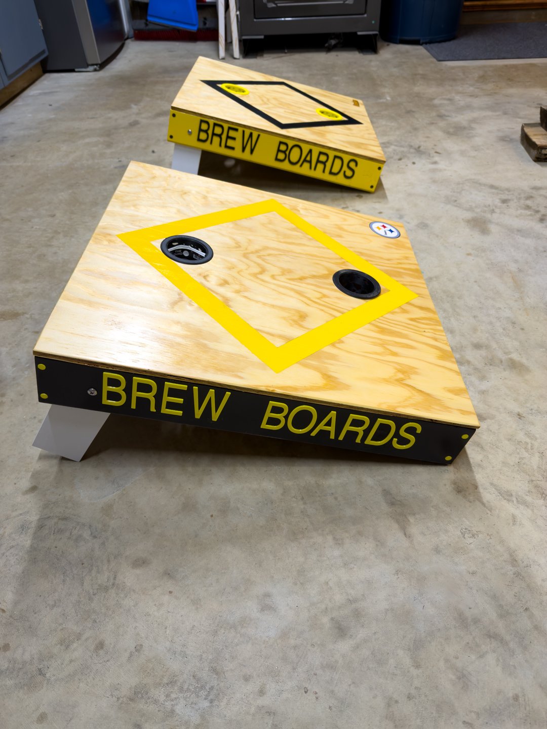 Two plywood tabletops engraved with BREW BOARDS, routed diamond outlines, and cup holders, on a shop floor