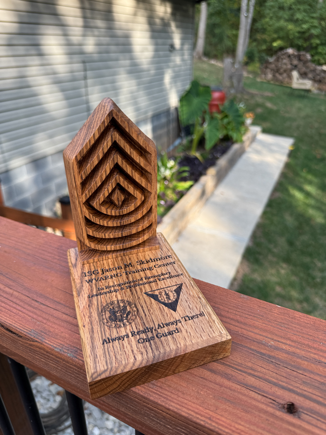 A laser-engraved wooden award plaque with chevrons and text standing on an outdoor deck railing