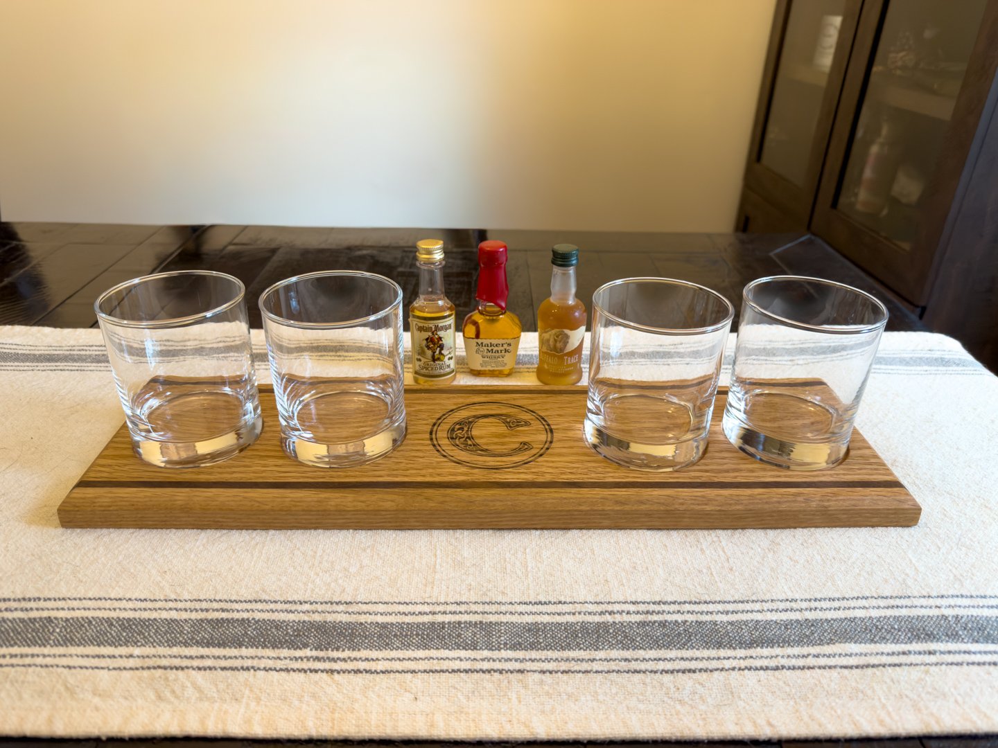 A light oak whiskey flight board with four empty glass holders, showing the engraved monogram in the center.